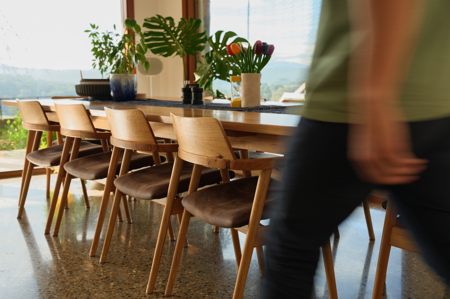 Timber dining chairs around a long table in a sunlit home, with a person walking past