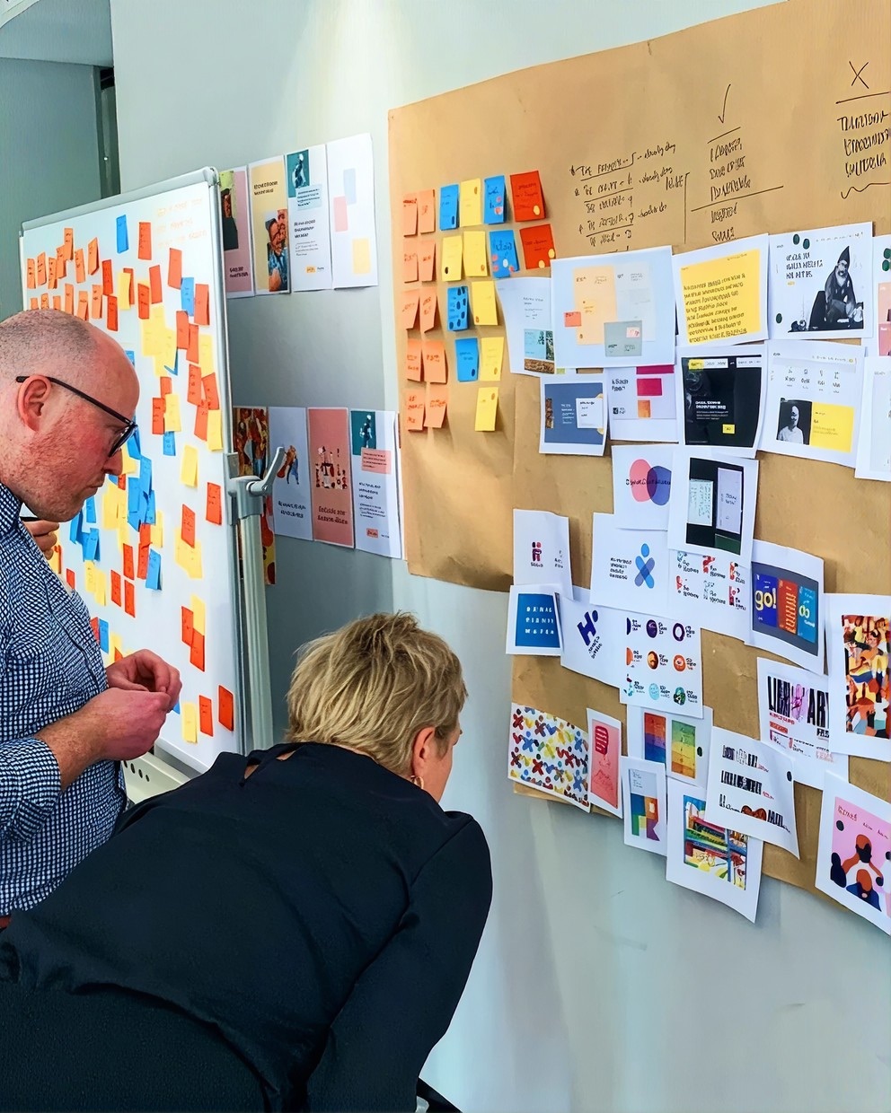 Two people studying a wall of sticky notes and pinned design references in a brand workshop