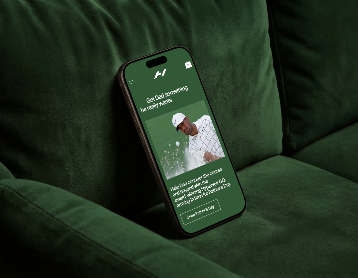 Phone on a green velvet sofa showing a Hyperice Father's Day page for the Hypervolt GO, with a golfer in a bunker