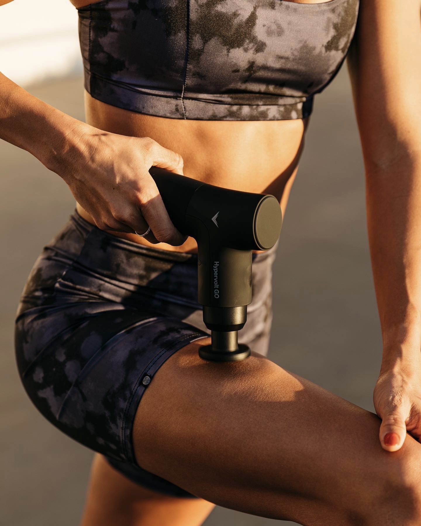 Woman using a black Hypervolt GO massage gun on her thigh in warm sunlight